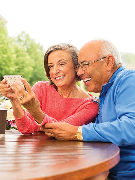 A couple sits together outside while smiling into a cellphone.