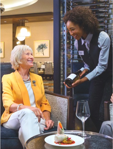 A woman in a yellow blazer is presented a bottle of wine by a Vi employee