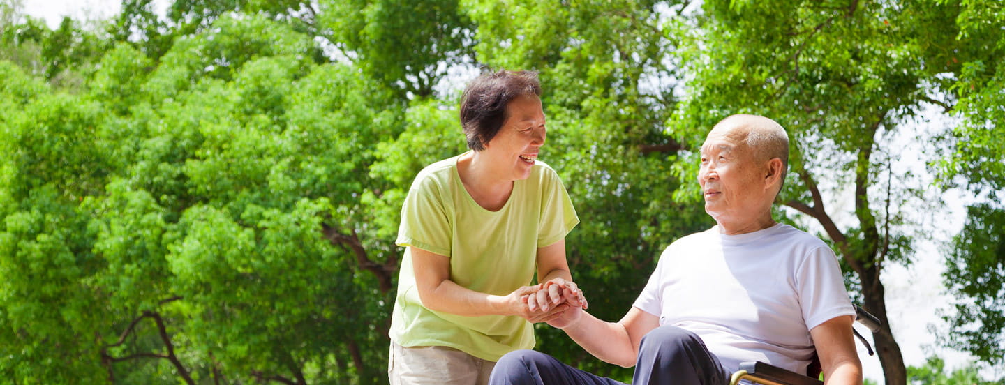 An Asian woman holds hands with an Asian man sitting in a wheelchair.