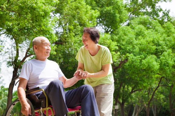 A man in a wheelchair looks into the distance while his wife smiles at him.