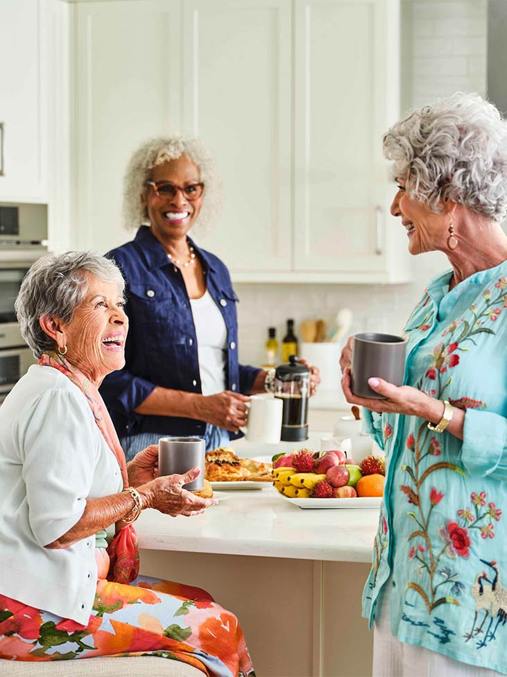 Three women hanging out in a kitchen with coffee.