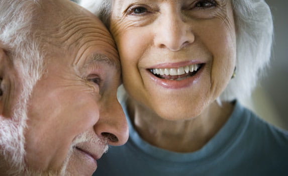 A woman smiles at the camera while a man leans his head against hers and looks off-camera.