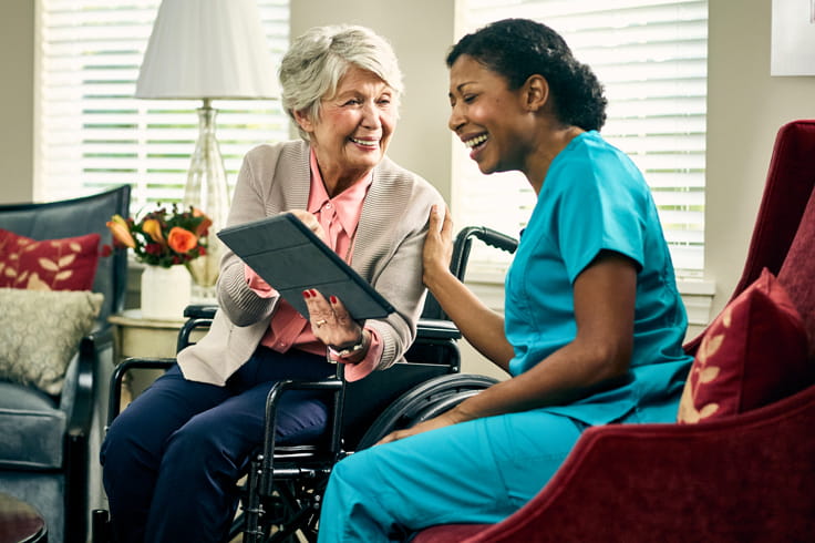 A woman and care professional sit side by side and look at a tablet screen.