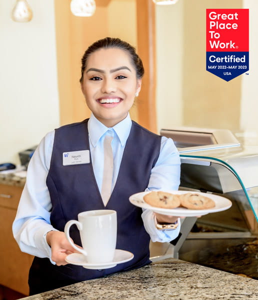A server holds cookies and coffee while smiling at the camera.