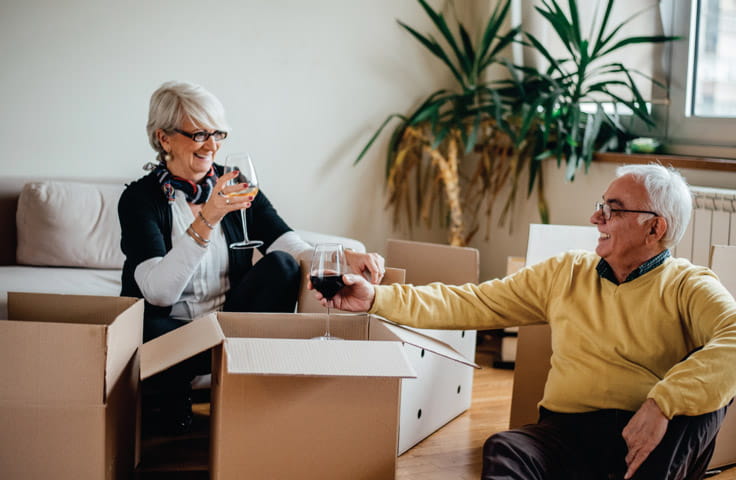 A man and woman toast with red wine while sitting among moving boxes.
