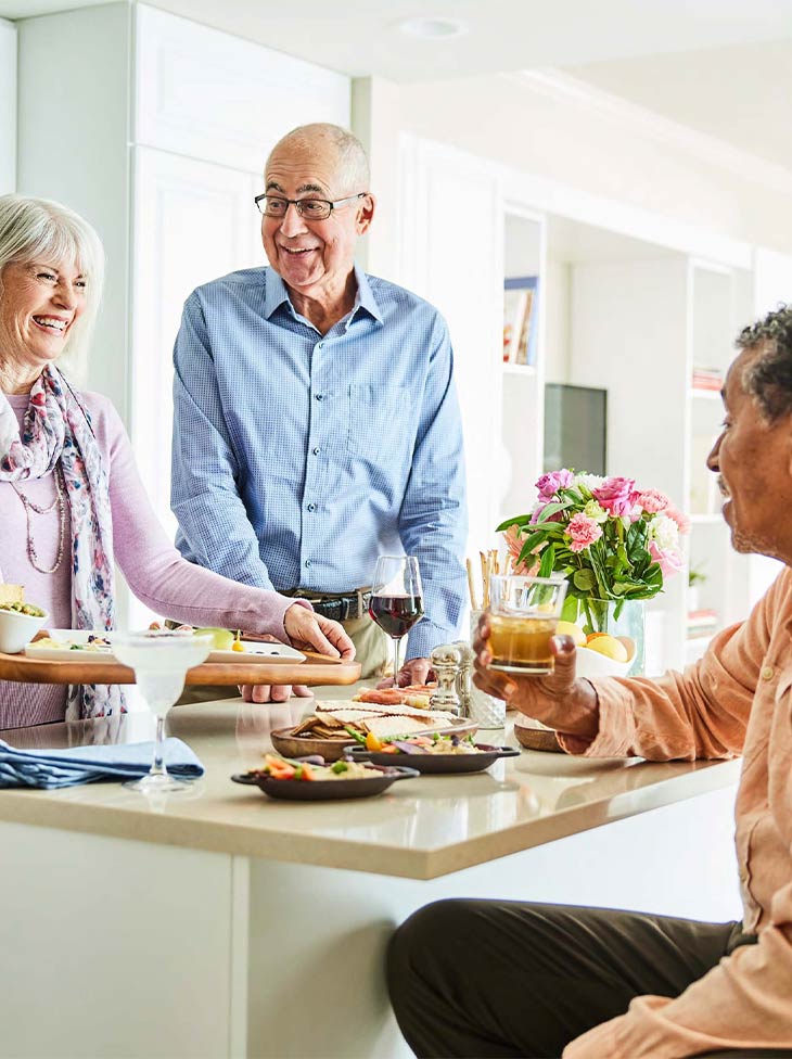 Three people hanging out in a kitchen with food.