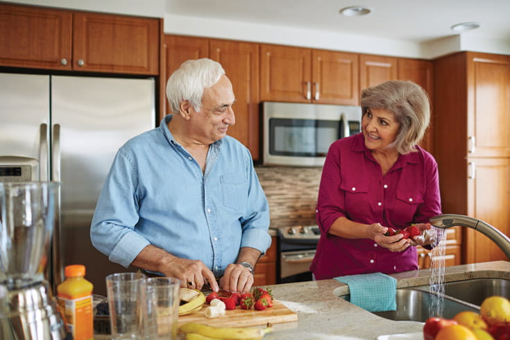 A couple smiles at each other while they prep food in the kitchen.