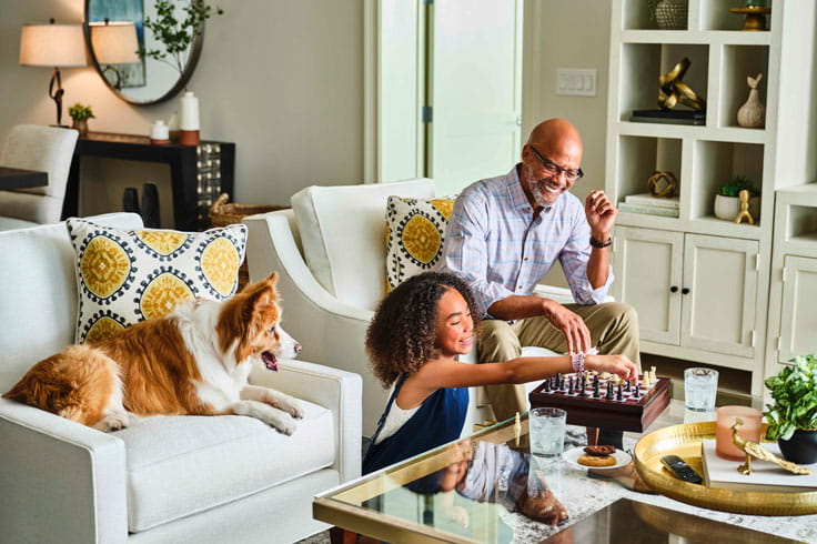 A older man and his grandchild play a game of chess while a dog looks on.