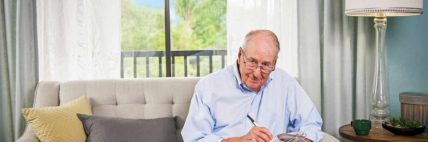A man looks over his glasses into the camera. He is sitting on a sofa and reading the paper.