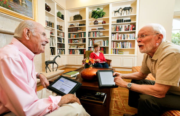 Two men discuss something in the library; a woman looks at her tablet in the background.