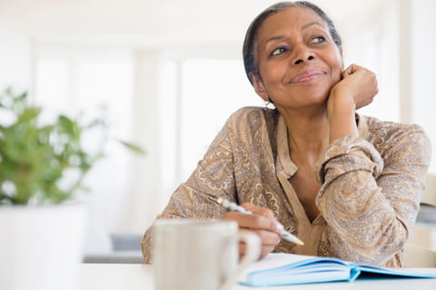 A woman looks into the distance with a pen poised over a notebook.